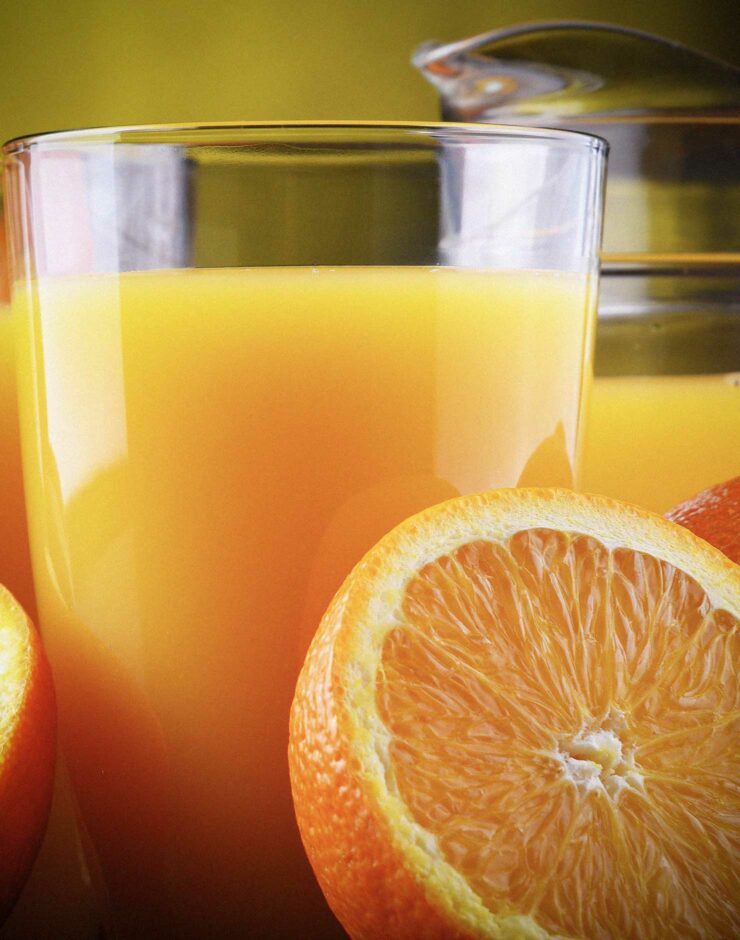 Close up of a glass of orange juice with half an orange in the foreground and a jug of orange juice in the background.