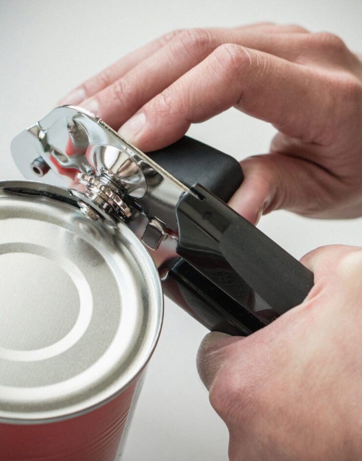A close-up of hands using a manual tin opener to open a metal tin. The opener grips the lid as one hand turns the handle and the other steadies the tin. The background is softly blurred.