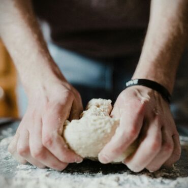 A person kneads a ball of dough on a floured surface, using both hands. Their sleeves are rolled up, and a black wristband is visible on one wrist.