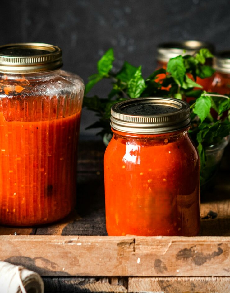Jars of homemade tomato sauce with metal lids are arranged on a rustic wooden tray, with fresh green herbs nearby and a dark, textured background behind them.