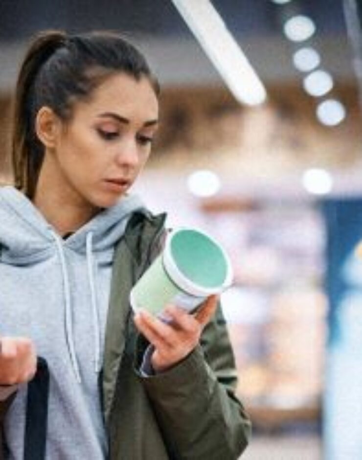 A woman in a green jacket looks closely at a product in a supermarket aisle while holding a shopping basket. She appears to be reading the label before making a decision.
