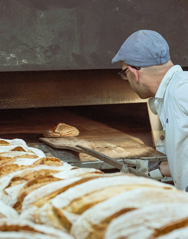 A baker wearing a blue cap and white shirt uses a peel to place or remove flour-dusted loaves of bread in a large industrial oven, with several fresh loaves lined up in the foreground.
