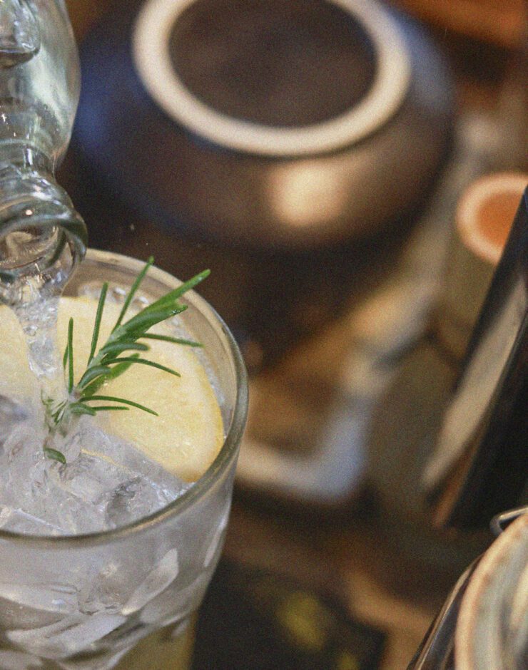 Pouring tonic into a glass of gin and ice, with a sprig of rosemary.