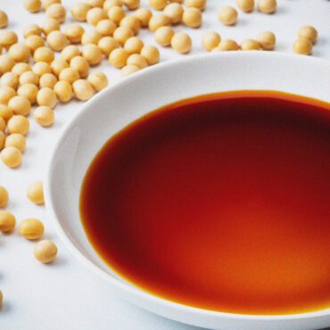 A white bowl filled with dark brown soy sauce sits on a white surface, surrounded by scattered whole soya beans.