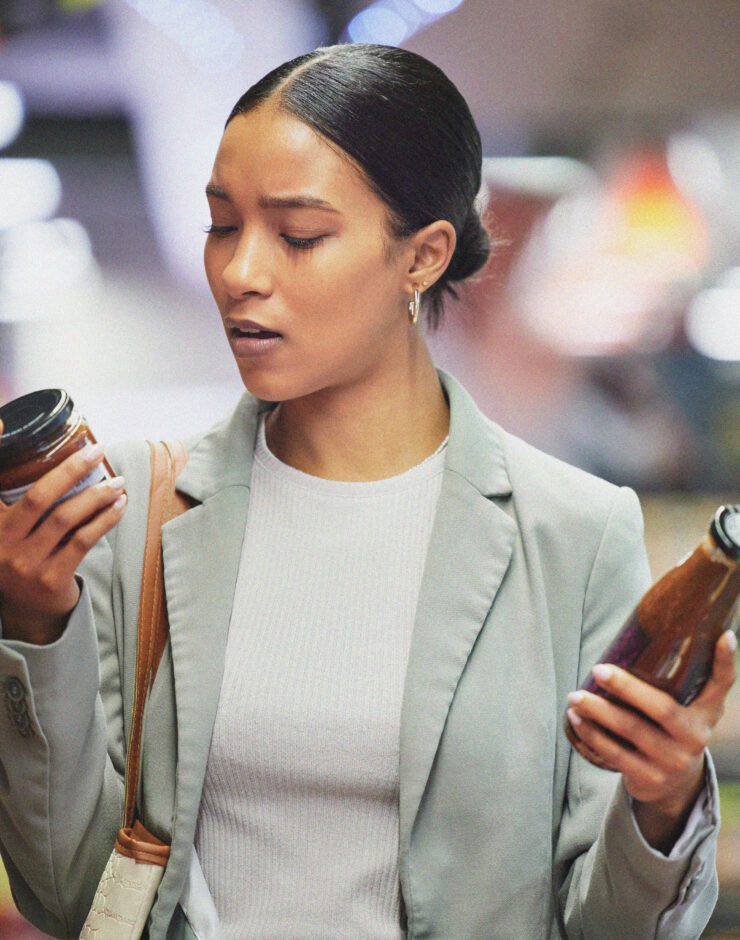 A woman in a shop holds a jar and a bottle, examining the labels intently for E numbers with a thoughtful expression. She is dressed in a light blazer and stands in an aisle lined with shelves of products in the background.