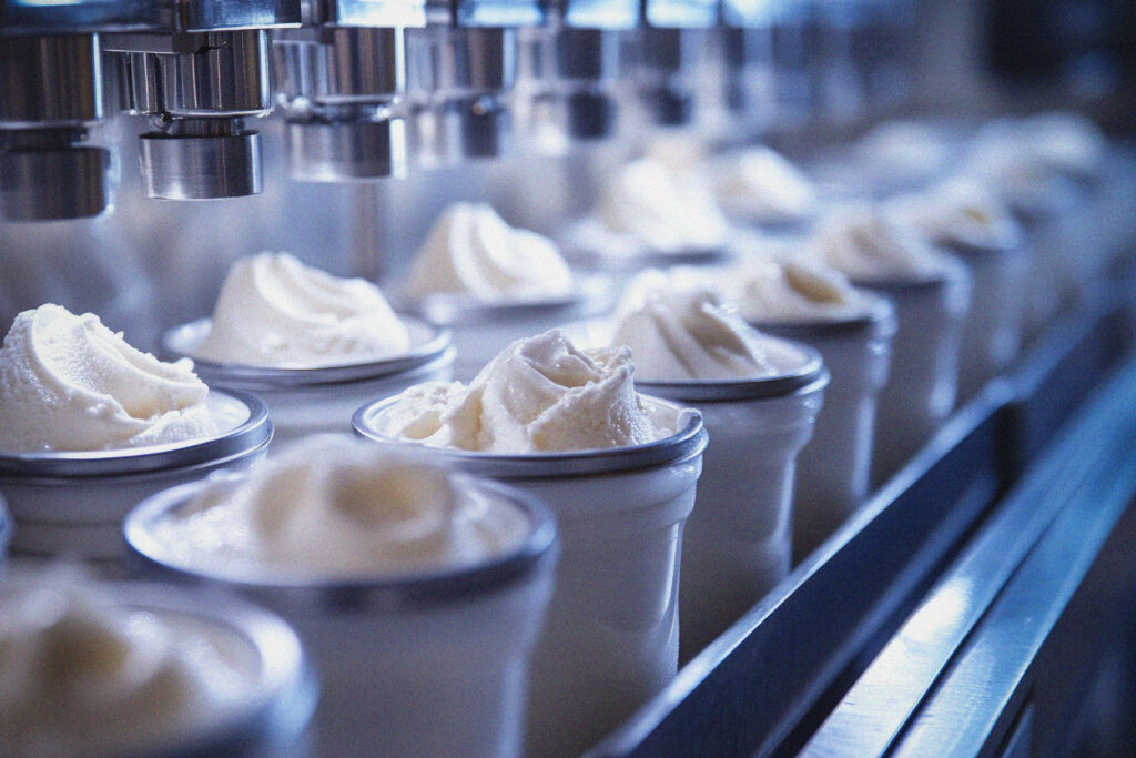 Cups being filled with soft-serve ice cream by an automated machine on a production line, with swirls of creamy white ice cream visible in each cup.