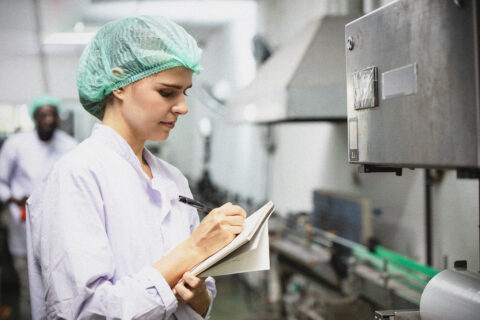 A woman in a lab coat and hairnet writes in a notebook whilst inspecting equipment in an industrial or laboratory setting. Conveyor belts and machinery are visible in the background.