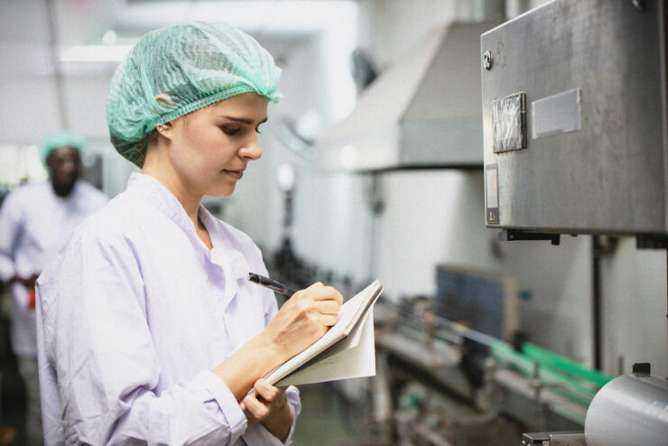 A woman in a lab coat and hairnet writes in a notebook whilst inspecting equipment in an industrial or laboratory setting. Conveyor belts and machinery are visible in the background.