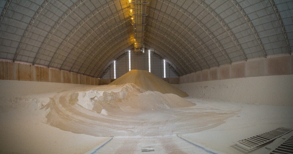 A large indoor storage facility with high arched ceilings, filled with mounds of white salt or sugar piled on the floor. Overhead lights illuminate the spacious warehouse interior.