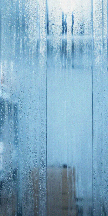 Foggy plastic strip curtains lead into a cold storage room with metal shelves holding cardboard boxes and containers; moisture and condensation are visible on the plastic strips.