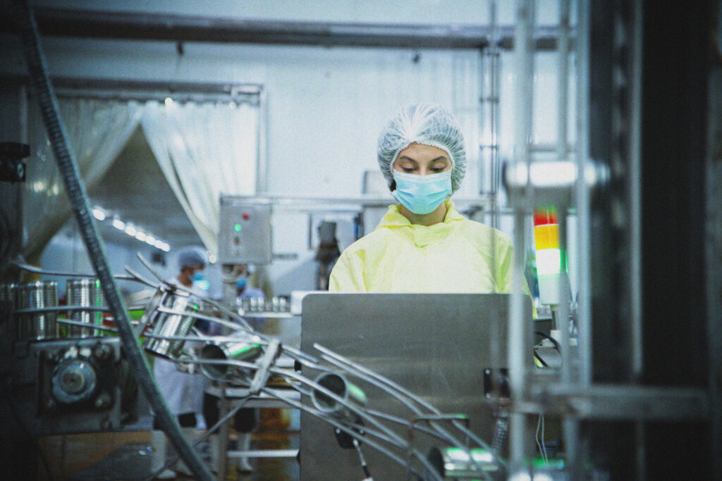 A person in protective clothing, hair net, and face mask operates machinery in a factory or laboratory setting, with another worker visible in the background.