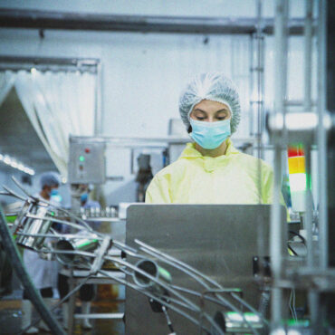 A person in protective clothing, hair net, and face mask operates machinery in a factory or laboratory setting, with another worker visible in the background.