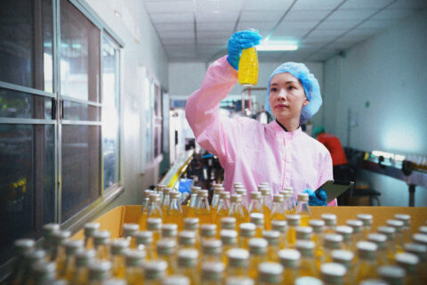 A person in a pink lab coat, blue hair net, and gloves inspects a yellow liquid in a bottle inside a factory with many similar bottles in the foreground.
