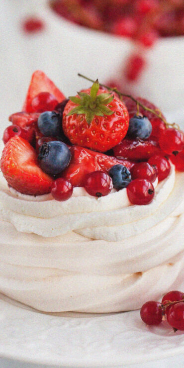 A pavlova, made with whipped egg whites and topped with strawberries, blueberries, and redcurrants, sits on a white plate beside a cup of coffee. A cluster of redcurrants rests on the plate, with a white teapot and a bowl of berries in the background.