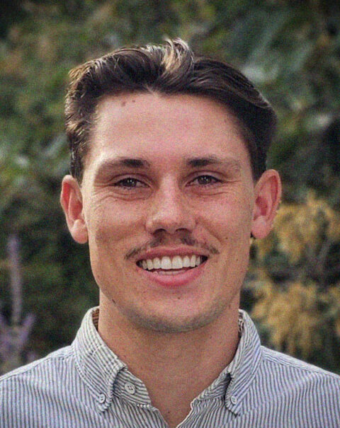 A young man with short brown hair and a moustache smiles at the camera, dressed in a striped button-up shirt. The blurred greenery in the background hints at his role as an Ingredients Distributor, connecting nature’s bounty to businesses with a friendly, professional presence.