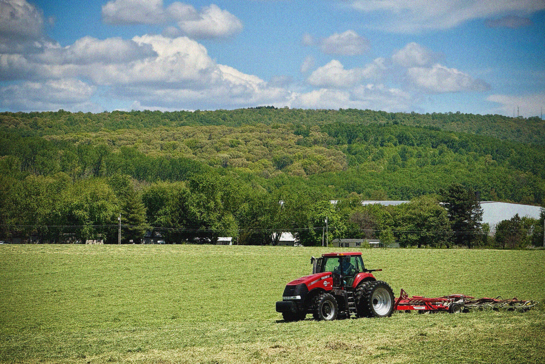 A red tractor pulls farming equipment across a large green field with dense trees and hills in the background under a partly cloudy sky.